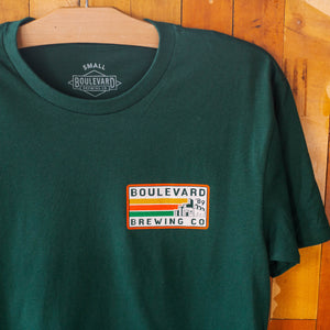 the front of a green t-shirt with a small logo that says Boulevard Brewing Co. '89 with a picture of the brewery and yellow, orange, and green stripes in a rectangle.