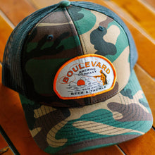 Load image into Gallery viewer, A camo trucker cap with a patch depicting a fisherman, that says, &quot;Boulevard Brewing Company, Since 1989, Beer &amp; Tackle&quot;, at an angled view.
