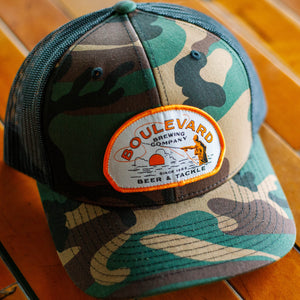 A camo trucker cap with a patch depicting a fisherman, that says, "Boulevard Brewing Company, Since 1989, Beer & Tackle", at an angled view.