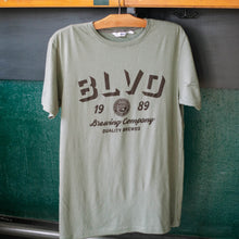 Load image into Gallery viewer, A sage green t-shirt hanging up in bright daylight.
