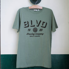 Load image into Gallery viewer, A sage green t-shirt hanging up.
