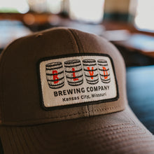 Load image into Gallery viewer, A sideview of a brown trucker cap with a white patch, spelling BLVD out in barrels.
