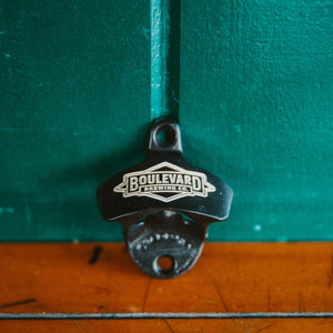 A metal wall mounted bottle opener with an etched diamond logo, leaning against a wall (front view)