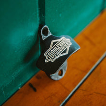 Load image into Gallery viewer, A metal wall mounted bottle opener with an etched diamond logo, leaning against a wall (side view)
