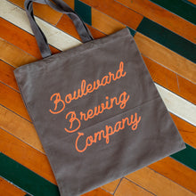 Load image into Gallery viewer, A grey-green tote bag with orange script lettering, laying on a wooden floor.
