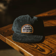 Load image into Gallery viewer, A black corduroy cap with a white and gold patch, on a wooden table.
