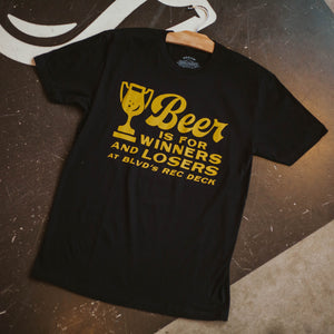 A black t-shirt, laying on the shuffleboard lanes,  with gold font, that says, "Beer is for Winners and Losers at BLVD's Rec Deck" with a trophy made out a beer glass.