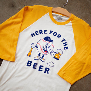 A yellow and white raglan tee with a baseball character carrying a beer and baseball bat that says, Here for the Beer, laying on the floor.