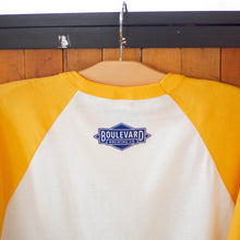 Load image into Gallery viewer, The back of a yellow and white raglan tee with a blue boulevard diamond logo near the top of the shirt.
