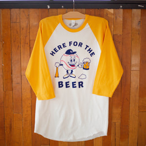 A yellow and white raglan tee with a baseball character carrying a beer and baseball bat that says, Here for the Beer, hanging up.
