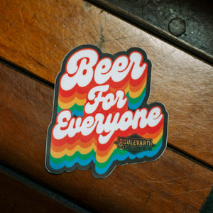 A rainbow sticker that says, "Beer for Everyone" against a wood floor.