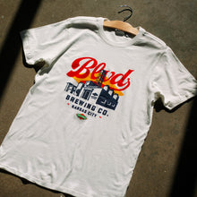 Load image into Gallery viewer, A white t-shirt with a brewery logo, laying on the ground.
