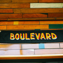 Load image into Gallery viewer, A black rectangular pennant that says, &quot;BOULEVARD&quot;, in colorful lettering laying on a wooden bench.
