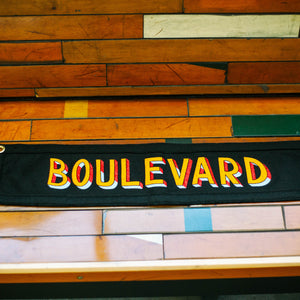 A black rectangular pennant that says, "BOULEVARD", in colorful lettering laying on a wooden bench.