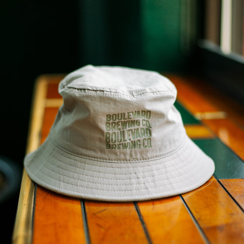 A tan bucket hat made of moisture-wicking material with green wavy lettering.