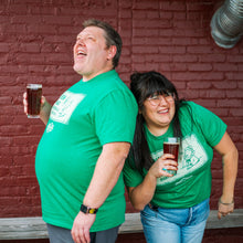 Load image into Gallery viewer, Two people wearing green shirts with a logo, standing against a brick wall.
