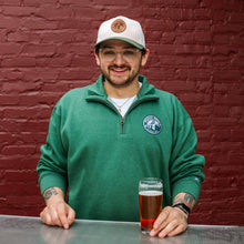 Load image into Gallery viewer, Man wearing a green sweater and cap with a logo, holding a glass of beer against a brick wall.
