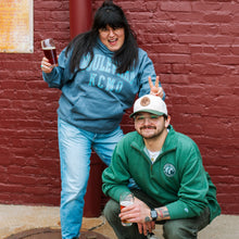 Load image into Gallery viewer, Two people posing against a brick wall, one holding a beer and the other wearing a green jacket with a logo.
