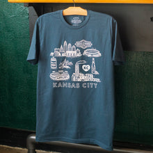 Load image into Gallery viewer, Blue t-shirt with white graphic design of Kansas City landmarks on a hanger against a green wall.
