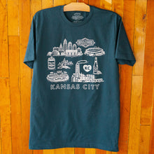 Load image into Gallery viewer, Blue t-shirt with white graphic design of Kansas City landmarks on a wooden surface
