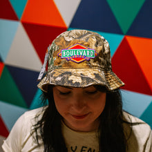 Load image into Gallery viewer, Person wearing a camouflage hat with Boulevard Brewing Co logo against a colorful geometric background
