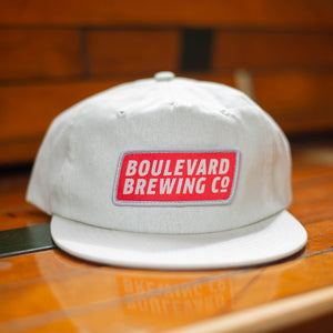 A natural colored flat-brim cap with a rectangular patch logo on a wooden bench.