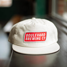 Load image into Gallery viewer, A natural colored flat-brim cap with a rectangular patch logo.
