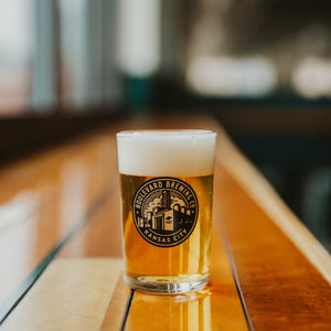 A small glass with a brewery logo on it filled with beer on a wooden table