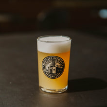 Load image into Gallery viewer, A small glass with a brewery logo on it filled with beer on a dark background
