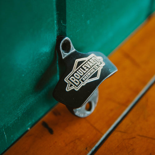A metal wall mounted bottle opener with an etched diamond logo, leaning against a wall (side view)