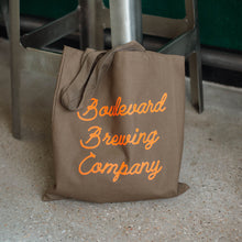 Load image into Gallery viewer, A partially full grey-green tote bag with orange script lettering, resting against the legs of a barstool.
