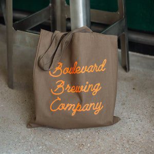 A partially full grey-green tote bag with orange script lettering, resting against the legs of a barstool.