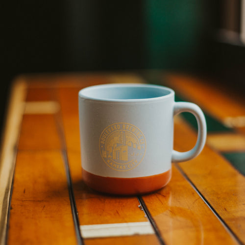 A baby blue ceramic mug with a terracotta bottom trim with the brewery logo