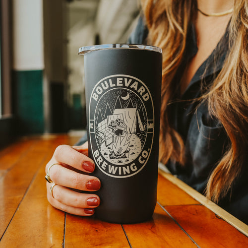 A woman's hand holding a black travel tumbler with a boulevard logo featuring a campsite