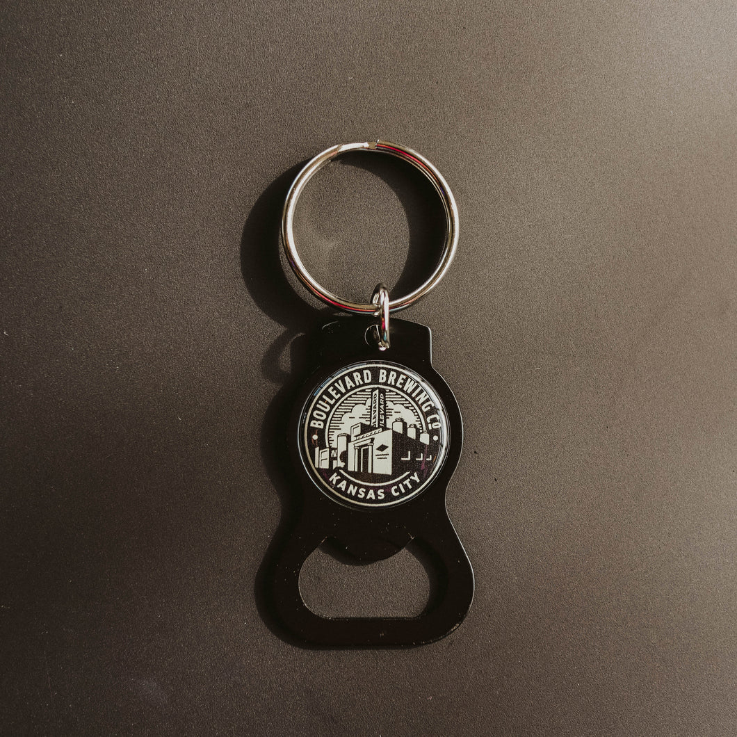 A black circular keycahin featurin the brewery logo and a bottle opener on the bottom